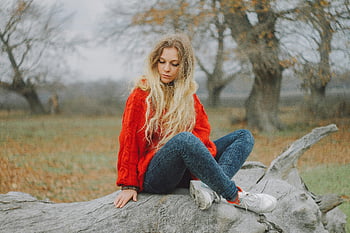 Frau im roten Pullover, auf dem Protokoll sitzen, blondes Haar, Outdoor-Szene, Jeans, Turnschuhe, Natur Hintergrund, Herbsteinstellung, Lässige Kleidung, Entspannte Pose, Baumstamm