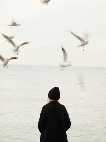 blond woman, black coat, seashore, seagulls flying, woman in hat, sea, flying seagulls, seashore scene, woman observing, seagulls, coastal scene