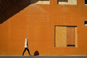 person walking, sidewalk, vibrant orange wall, casting shadow, city scene, urban walk, street photography, architectural photography, outdoor scene, modern city, person in motion