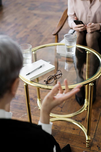 therapy session, woman in black shirt, seated at table, glass table, notebooks, water glasses, two adults, therapy, conversation, professional setting, counseling