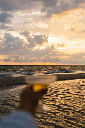hand holding glass, sea shore, sunset, blurry hand, seaside sunset, beach sunset, ocean view, golden hour, coastal scene, relaxing moment, drink at sunset