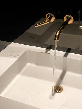 water flowing, bronze faucet, modern washbasin, gold faucet, dimly lit bathroom, elegant bathroom, white washbasin, sleek faucet, water stream, bathroom setting, bronze colored faucet