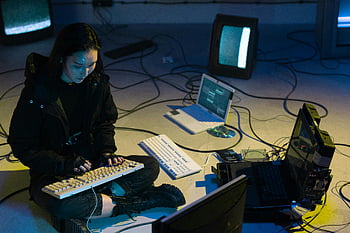 Young Asian woman, computer hacking, dimly lit room, technologically equipped, typing on keyboard, laptop, monitors, cables, hacking setup, cyber security, digital crime