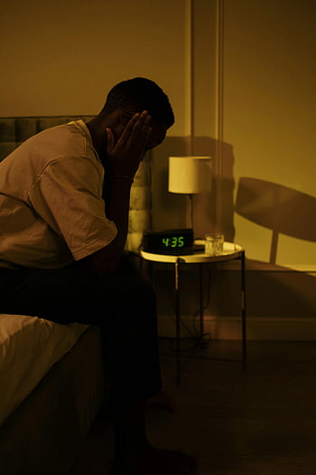 Uomo seduto sul letto, mattina, camera da letto debolmente illuminata, uomo premuroso, bordo del letto, comodino, sveglia, lampada, mattina presto, Scena della camera da letto, umore contemplativo