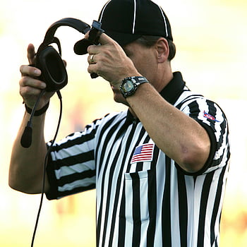 football referee, black fitted cap, black corded headset, adjusting headset, outdoor game, referee uniform, striped shirt, American flag patch, sports official, game official, sports referee