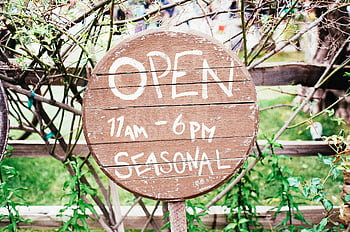brown and white signage, round wooden sign, open sign, 11am-6pm, seasonal opening, greenery background, outdoor sign, business hours, rustic sign, garden sign, seasonal business