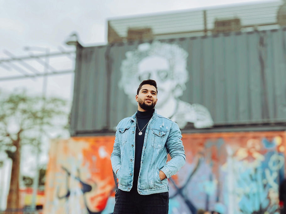 kék farmerdzsekije, gombos kabát, divatos fiatalember, graffiti falfestmény, kültéri divat, magabiztos póz, utcai stílus, városi divat, farmer divat, színes graffiti, szabadtéri portré