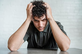 young man, upset, sitting, holding head, hands, stress, worry, indoors, curly hair, black shirt, table