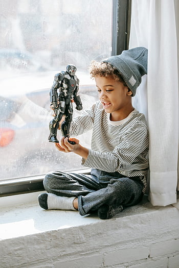 positive black boy, joyful child, curly hair, toy robot, sunny window, indoors, playing, beanie hat, striped shirt, gray pants, socks