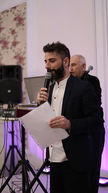 man in black suit, speaking into microphone, indoor event, holding paper, formal attire, public speaking, event speaker, microphone use, professional setting, event photography, indoor venue