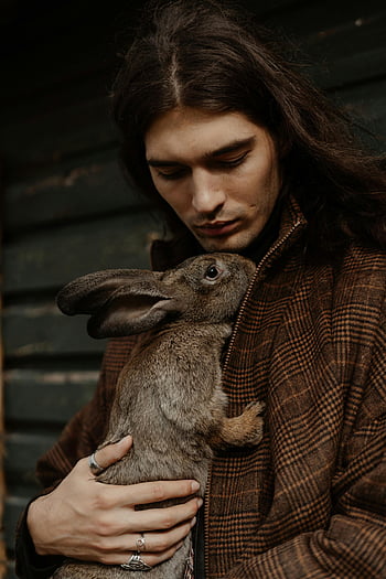 man in brown coat, holding rabbit, young man, wavy hair, cozy outdoor setting, brown rabbit, outdoor photo, man with rabbit, brown coat, wavy-haired man, cozy setting