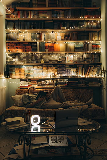 운동복 바지를 입은 남자, 소파에 누워 있습니다, 책장, 끈 조명, 따뜻한 분위기, 축제 분위기, 실내 공간, 소파에서 책 읽기, 아늑한 설정, 가정 도서관, 편안한 독서