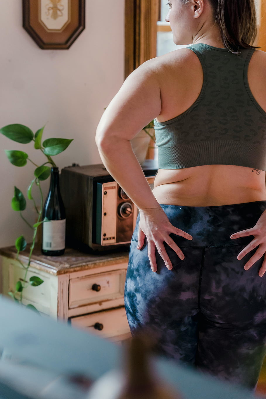 woman checking hips, confident woman, active wear, body positivity, indoors, sports bra, tie-dye leggings, home decor, vintage radio, plant, tattoo
