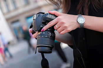 woman holding camera, black DSLR camera, outdoor photography, hands adjusting camera, photography themes, woman photographer, DSLR device, outdoor photo session, camera focus, street photography, woman with watch