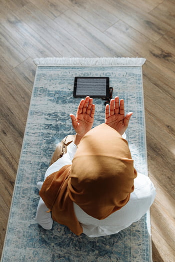 woman praying, hijab, blue prayer rug, tablet, peaceful indoor setting, high-angle shot, kneeling, spiritual practice, religious devotion, indoor prayer, digital Quran