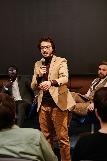 ビジネス会議、 スピーカー、 視聴者のエンゲージメント、 インタラクティブな議論、 茶色のスーツジャケット、 マイクロフォン、 プロの服装、 パネル・ディスカッション、 フォーマルイベント、 人前で話す、 会議室