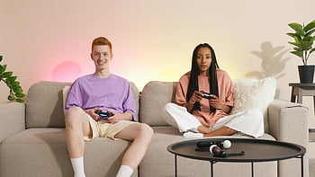Pár hrající videohry, sedí na gauči, uvolněné hraní v interiéru, Ovládače videoher, útulný obývací pokoj, příležitostné herní sezení, pohodlný gauč, Vnitřní zábava, herní pár, domácí herní sestava, uvolněná atmosféra