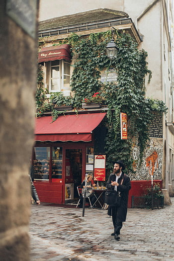 man walking, cozy ivy-covered café, narrow cobblestone street, red awning, ivy-covered building, outdoor seating, urban scene, street photography, European architecture, city life, man in hat