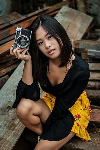 woman in black dress, holding camera, vintage camera, rustic outdoor setting, stylish young woman, posing, outdoor photography, black dress, vintage photography, rustic background, yellow skirt