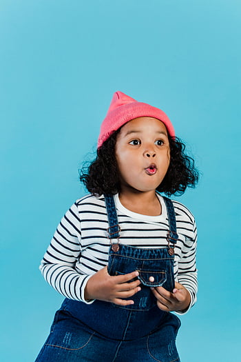 Sorpresa ragazza nera, muro blu, bel bimbo, capelli ricci, Outfit a strisce, cappello rosa, espressione sorpresa, sfondo blu, ragazza, salopette di jeans, camicia a righe