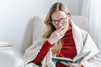 sleepy student, young female, yawning, homework assignment, cozy blanket, indoor reading, book, glasses, home study, relaxed learning, comfortable environment