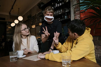 man in yellow hoodie, ordering, sign language, waitress, nonverbal gestures, cozy café, masked waitress, woman, man, table, coffee cups