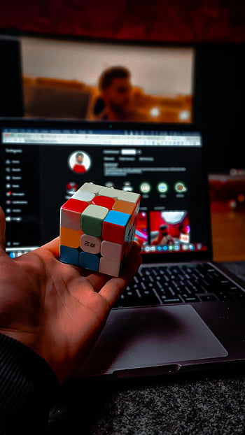 Rubik's Cube, Studio Laptop, hand holding, blurred screen, indoors, puzzle solving, cube mysteries, laptop screen, indoor setting, Rubik's Cube tutorial, computer screen