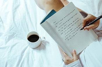 woman writing, coffee cup, notebook, cozy indoor, bed, writing in notebook, cup of coffee, cozy background, indoor setting, writing activity, white bed sheets