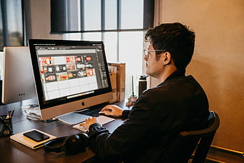 man working on computer, graphic design work, modern office setting, natural lighting, computer screen, desk setup, headphones, workspace, professional environment, indoor office, man using computer