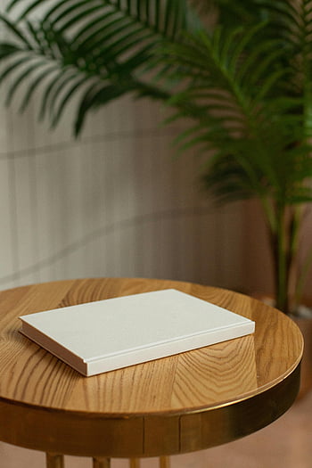 faasztal, Üres keményen elfojtott könyv, minimalista fehér notebook, kerek faasztal, homályos növényi háttér, minimalista design, fehér notebook, fa felület, beltéri környezet, növényi dekoráció, egyszerű összetétel