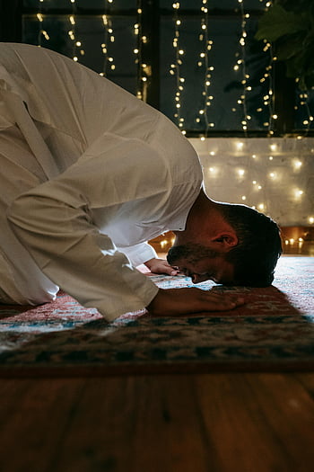 man in white thobe, bowing down, red and blue rug, traditional attire, prayer, illuminated string lights, indoors, cultural attire, spiritual moment, prayer rug, string lights
