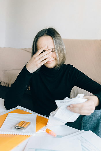woman in black shirt, sitting at table, overwhelmed by bills, holding receipts, financial stress, indoor setting, young woman, long sleeve shirt, desk clutter, financial management, emotional distress