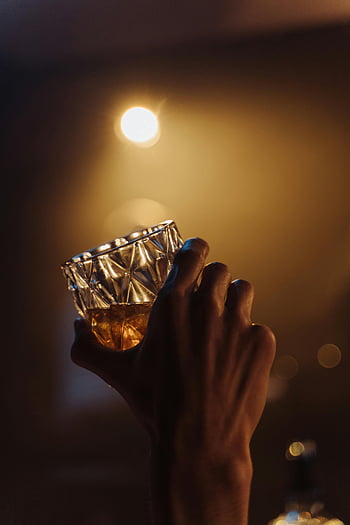Ruční držení whisky, dekorativní sklo, teplé osvětlení, Sofistikovaná atmosféra, detailní záběr, sklenice na whisky, osvětlená ruka, elegantní nápoj, Útulná atmosféra, Amber kapalina, rafinované nastavení