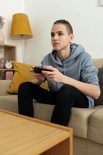 dospívající chlapec, šedá mikina s kapucí, černé kalhoty, sedí na gauči, hraní videoher, vážný výraz, Útulné vnitřní nastavení, žlutý polštář, svítilna, herního řadiče, obývací pokoj