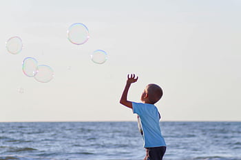 boy playing bubbles, white t-shirt, body of water, grey sky, daytime, joyful child, ocean, summer fun, beach, bubbles, sunny day
