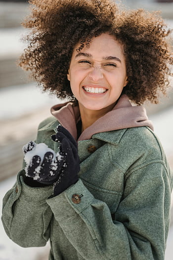 donna etnica, palla di neve, donna sorridente, capelli ricci, inverno all'aperto, godersi l'inverno, la neve, guanti, cappotto verde, felpa rosa, divertimento all'aria aperta