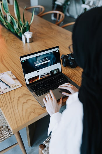 woman in hijab, working on laptop, wooden table, camera nearby, young woman, using laptop, hijab, laptop, camera, workspace
