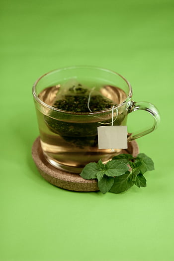 green tea, mint leaves, cup holder, hot herbal tea, cork coaster, bright green background, tea bag, clear glass mug, refreshing drink, healthy beverage, natural tea