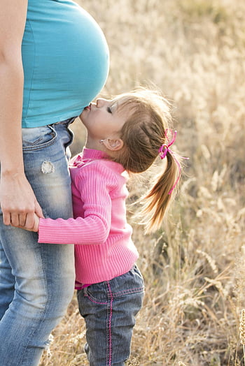 girl in pink sweater, grey jeans, kissing tummy, pregnant woman, blue shirt, blue denim jeans, family affection, outdoors, young girl, mother's belly, love