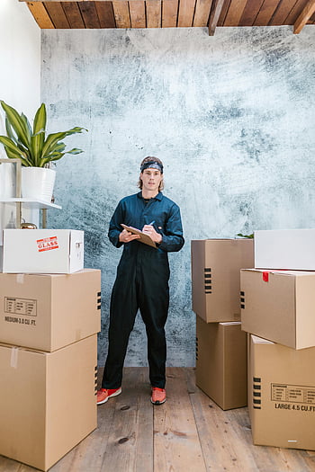 pria berpakaian overall, sepatu karet, memegang clipboard, kotak kardus, Penggerak profesional, mengatur paket, hari yang bergerak, Pengaturan dalam ruangan, lantai kayu, menanam, latar belakang dinding