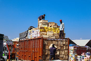 loading cargo truck, wooden boxes, three men, clear blue sky, Sopore, cargo transportation, manual labor, outdoor work, truck loading, industrial scene, workers