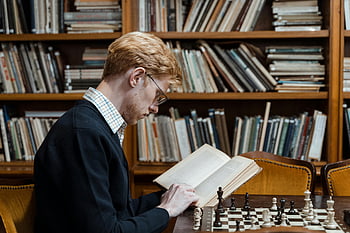 man reading book, chess game, library setting, side view, bookshelf, indoor scene, focused reader, intellectual activity, chess pieces, study environment, quiet reading