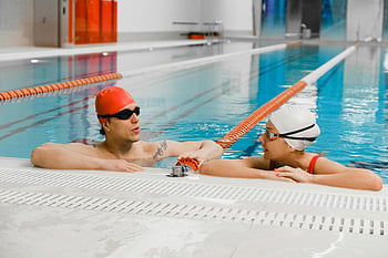 man and woman talking, indoor pool, swimmers with caps, swimmers with goggles, conversation at pool edge, indoor swimming pool, poolside conversation, swimmer interaction, poolside swimmers, swimming caps, swimming goggles