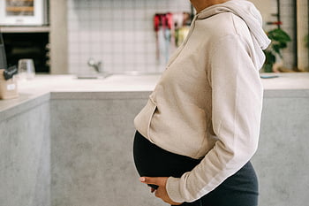 schwangere Frau, Babybauch, Casual Hoodie, Bauch halten, drinnen, Kücheneinstellung, Umstandsmode, Schwangerschaftsstil, werdende Mutter, Heimumgebung, Sanfte Berührung