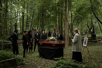 funeral, forested cemetery, people mourning, priest present, solemn gathering, honoring departed, cemetery scene, group gathering, outdoor funeral, wooded cemetery, memorial service