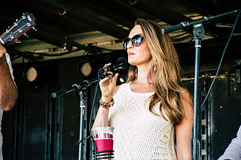 woman singing, microphone, sunglasses, outdoor concert, stage performance, confident singer, stylish woman, live music, white top, long hair, microphone stand