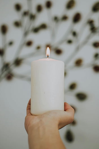 hand holding candle, lit white candle, peaceful ambiance, blurred backdrop, artistic background, white pillar candle, hand holding lit candle, candlelight, soft lighting, serene atmosphere, minimalist decor