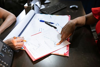 person pointing, white printer paper, discussing graphs, data analysis, desk collaboration, writing notes, office work, team discussion, business meeting, graph analysis, data visualization