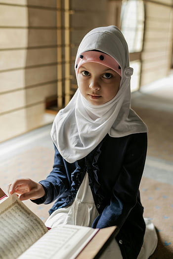 Pige i hijab, læser Koranen, indendørs portræt, ung pige, Hvid hijab, blå og hvid skjorte, fredeligt øjeblik, Kulturel scene, indendørs indstilling, religiøs aktivitet, Koranundersøgelsen