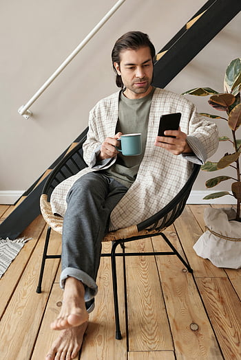 man sitting, mug, smartphone, indoor, comfortable attire, relaxing, coffee mug, smartphone use, casual setting, wooden floor, modern chair